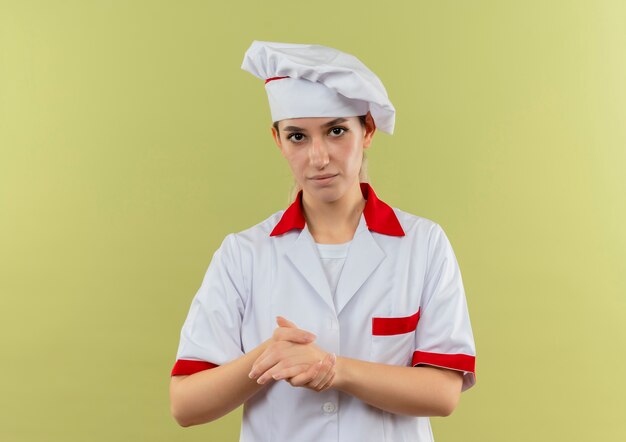 Cocinero bastante joven confiado en uniforme del cocinero que mantiene las manos juntas aisladas en la pared verde