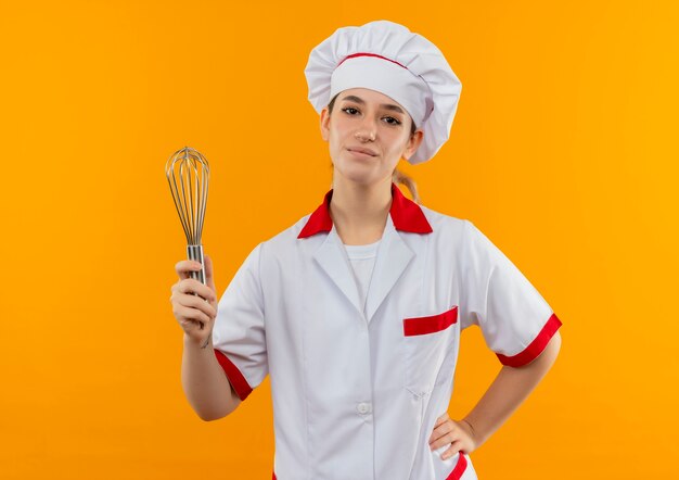 Cocinero bastante joven confiado en uniforme del cocinero que sostiene el batidor con la mano en la cintura aislada en la pared naranja