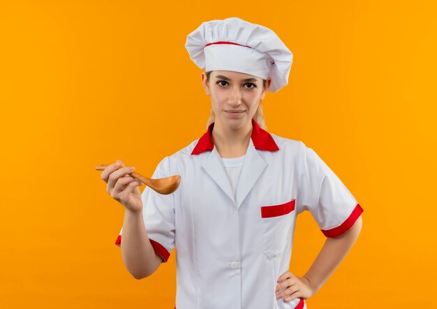 Cocinero bastante joven confiado en uniforme del cocinero que sostiene la cuchara que pone la mano en la cintura aislada en la pared naranja