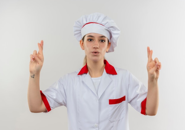 Cocinero bastante joven impresionado en uniforme del cocinero que hace gesto de los dedos cruzados aislado en la pared blanca