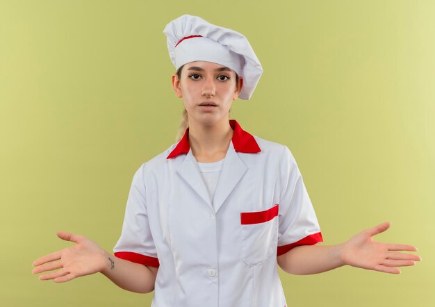 Cocinero bastante joven impresionado en uniforme del cocinero que muestra las manos vacías aisladas en la pared verde