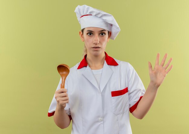 Cocinero bastante joven impresionado en uniforme del cocinero que sostiene la cuchara que muestra la mano vacía aislada en la pared verde