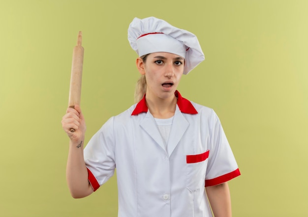 Cocinero bastante joven impresionado en uniforme del cocinero que sostiene el rodillo aislado en la pared verde