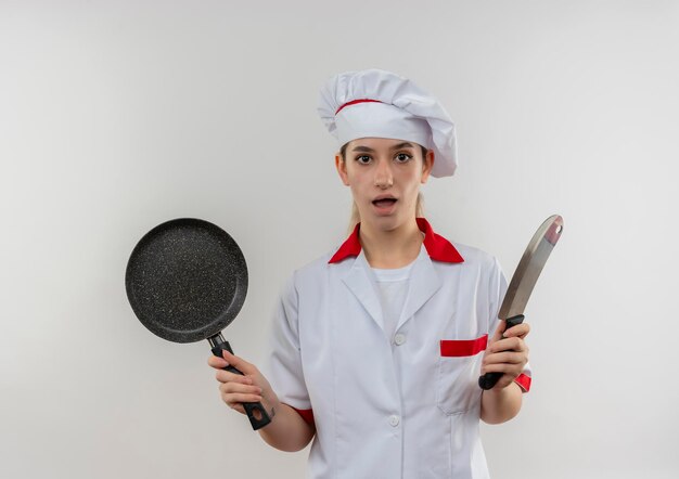 Cocinero bastante joven impresionado en uniforme del cocinero que sostiene la sartén y el cuchillo aislado en la pared blanca