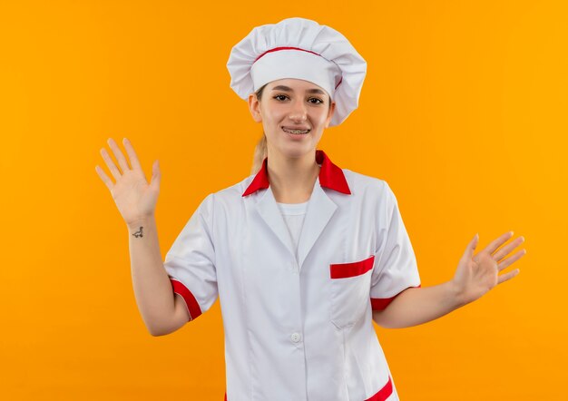 Cocinero bastante joven sonriente en uniforme del cocinero con los apoyos dentales que muestran las manos vacías aisladas en el espacio anaranjado