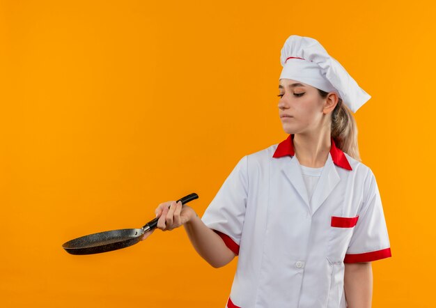 Cocinero bastante joven en uniforme del cocinero que sostiene y que mira la sartén