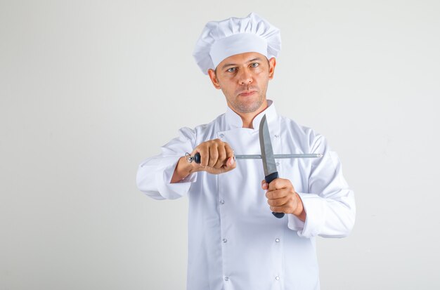 Cocinero chef masculino con cuchillos de cocina en uniforme y sombrero y mirando confiado