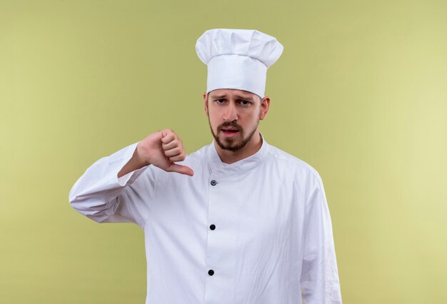 Cocinero de chef masculino profesional autosatisfecho en uniforme blanco y sombrero de cocinero apuntando a sí mismo de pie sobre fondo verde