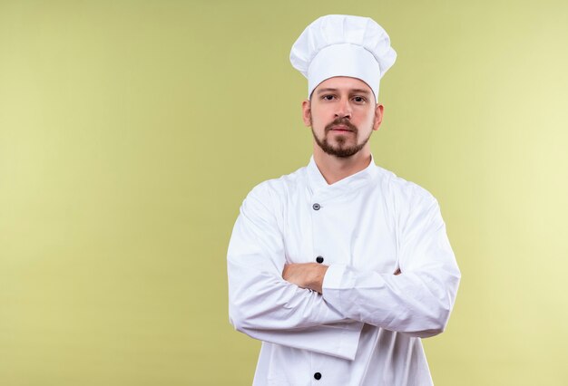 Cocinero chef masculino profesional satisfecho en uniforme blanco y sombrero de cocinero de pie con los brazos cruzados mirando confiado sobre fondo verde