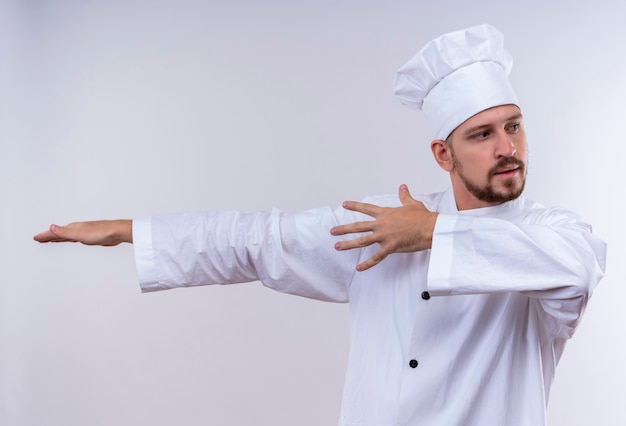 Cocinero de chef masculino profesional en uniforme blanco y sombrero de cocinero apuntando con los brazos y las manos al lado de pie sobre fondo blanco