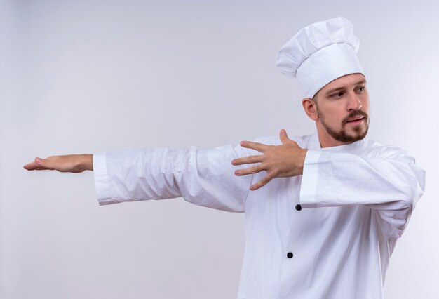 Cocinero de chef masculino profesional en uniforme blanco y sombrero de cocinero apuntando con los brazos y las manos al lado de pie sobre fondo blanco