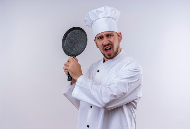 Cocinero de chef masculino profesional en uniforme blanco y sombrero de cocinero balanceando una sartén con expresión agresiva de pie sobre fondo blanco