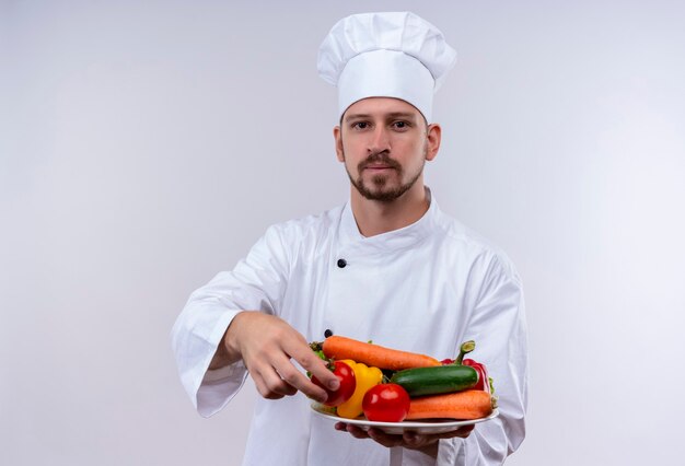 Cocinero de chef masculino profesional en uniforme blanco y sombrero de cocinero demostrando un plato con verduras mirando confiado de pie sobre fondo blanco