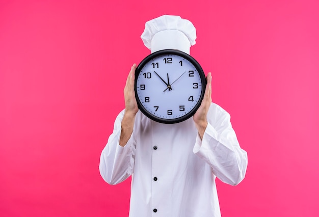 Cocinero de chef masculino profesional en uniforme blanco y sombrero de cocinero sosteniendo un gran reloj escondido detrás de él de pie sobre fondo rosa