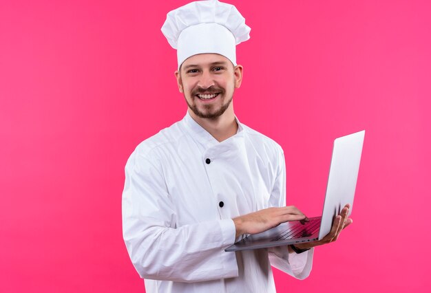 Cocinero de chef masculino profesional en uniforme blanco y sombrero de cocinero sosteniendo portátil mirando a la cámara sonriendo alegremente de pie sobre fondo rosa