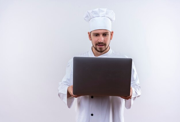 Cocinero de chef masculino profesional en uniforme blanco y sombrero de cocinero sosteniendo portátil mirándolo intrigado de pie sobre fondo blanco