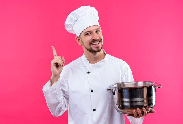 Cocinero de chef masculino profesional en uniforme blanco y sombrero de cocinero sosteniendo una sartén apuntando con el dedo hacia arriba sonriendo alegremente de pie sobre fondo rosa