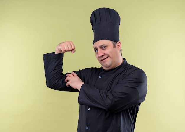 Cocinero chef masculino vistiendo uniforme negro y gorro de cocinero levantando el puño mostrando bíceps mirando confiados de pie sobre fondo verde