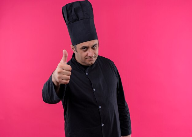 Cocinero chef masculino vistiendo uniforme negro y gorro de cocinero mirando a la cámara sonriendo mostrando los pulgares para arriba de pie sobre fondo rosa