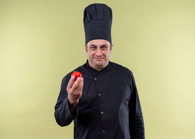 Cocinero chef masculino vistiendo uniforme negro y sombrero de cocinero mostrando tomate sonriendo mirando a la cámara de pie sobre fondo verde