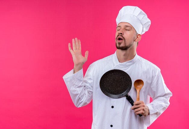 Cocinero cocinero masculino profesional molesto en uniforme blanco y sombrero del cocinero que sostiene la cacerola y la cuchara de madera que cierran los ojos que se colocan sobre fondo rosado