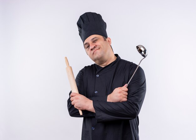 Cocinero cocinero masculino con uniforme negro y sombrero de cocinero sosteniendo la cuchara y el rodillo guiñando un ojo y sonriendo mirando a la cámara de pie sobre fondo blanco.