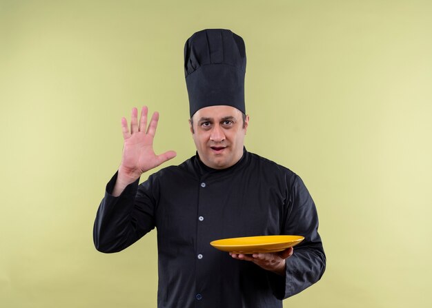 Cocinero cocinero masculino vistiendo uniforme negro y gorro de cocinero sosteniendo un plato vacío mostrando el brazo número cinco de pie sobre fondo verde