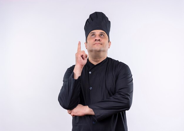 Cocinero cocinero masculino vistiendo uniforme negro y sombrero de cocinero apuntando con el dedo índice hacia arriba mirando hacia arriba de pie sobre fondo blanco.