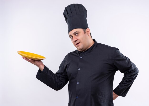 Cocinero cocinero masculino vistiendo uniforme negro y sombrero de cocinero demostrando plato mirando confiado de pie sobre fondo blanco.