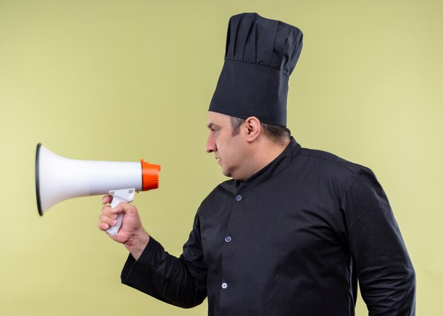 Cocinero cocinero masculino vistiendo uniforme negro y sombrero de cocinero mirando a un lado con cara seria sosteniendo megáfono de pie sobre fondo verde