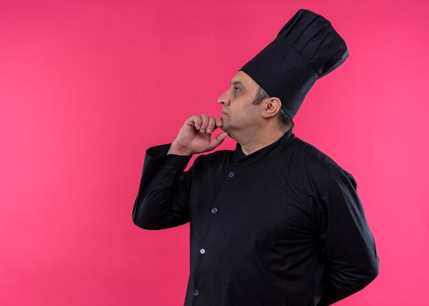 Cocinero cocinero masculino vistiendo uniforme negro y sombrero de cocinero mirando a un lado con la mano en la barbilla con expresión pensativa de pie sobre fondo rosa