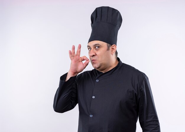 Cocinero cocinero masculino vistiendo uniforme negro y sombrero de cocinero que muestra el signo de pie delicioso sobre fondo blanco