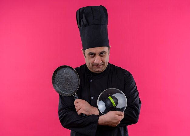 Cocinero cocinero masculino vistiendo uniforme negro y sombrero de cocinero sosteniendo una cacerola cruzando las manos mirando a la cámara con rostro serio de pie sobre fondo rosa