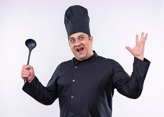 Cocinero cocinero masculino vistiendo uniforme negro y sombrero de cocinero sosteniendo la cuchara levantando el brazo mirando a la cámara con expresión agresiva de pie sobre fondo blanco.