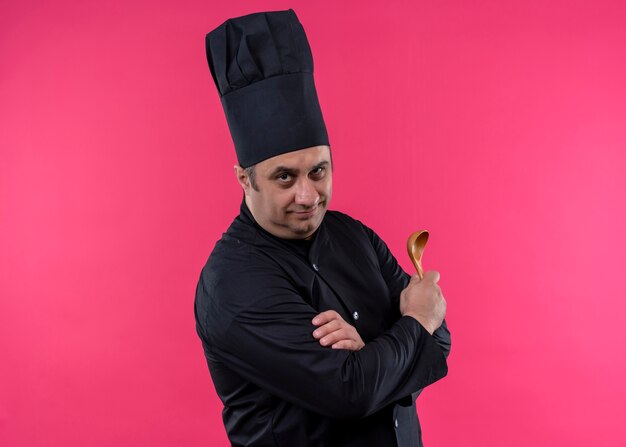 Cocinero cocinero masculino vistiendo uniforme negro y sombrero de cocinero sosteniendo una cuchara de madera mirando a la cámara con expresión de confianza de pie sobre fondo rosa