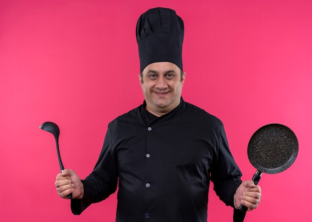 Cocinero cocinero masculino vistiendo uniforme negro y sombrero de cocinero sosteniendo pan y cucharón sonriendo confiado de pie sobre fondo de color rosa