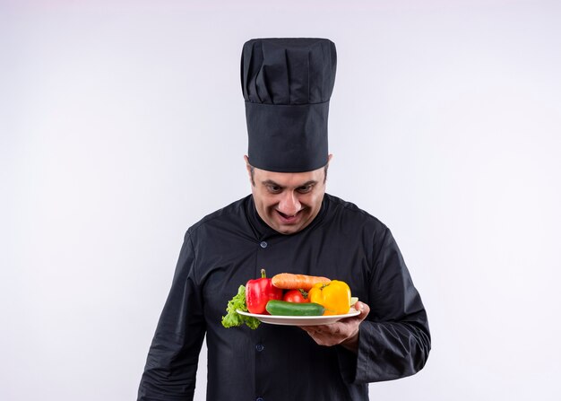 Cocinero cocinero masculino vistiendo uniforme negro y sombrero de cocinero sosteniendo la placa con verduras frescas mirando la placa con una sonrisa en la cara de pie sobre fondo blanco