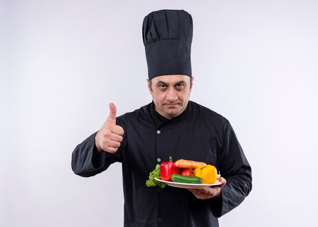 Cocinero cocinero masculino vistiendo uniforme negro y sombrero de cocinero sosteniendo la placa con verduras frescas mostrando los pulgares para arriba de pie sobre fondo blanco.