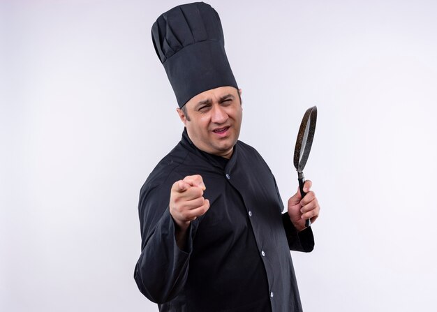 Cocinero cocinero masculino vistiendo uniforme negro y sombrero de cocinero sosteniendo una sartén apuntando con el dedo a la cámara de pie feliz y positivo sobre fondo blanco