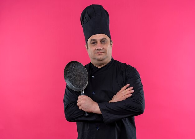 Cocinero cocinero masculino vistiendo uniforme negro y sombrero de cocinero sosteniendo una sartén mirando a la cámara con expresión de confianza de pie sobre fondo rosa