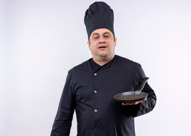 Cocinero cocinero masculino vistiendo uniforme negro y sombrero de cocinero sosteniendo una sartén con los ojos cerrados de pie sobre fondo blanco.