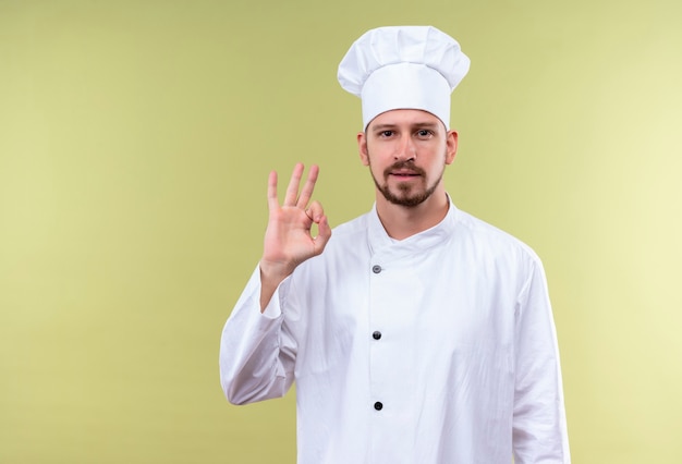 Cocinero cocinero profesional masculino en uniforme blanco y gorro de cocinero mostrando gesto ok mirando confiado de pie sobre fondo verde