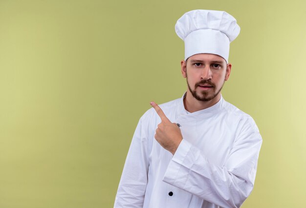 Cocinero cocinero profesional masculino en uniforme blanco y sombrero de cocinero apuntando con el dedo hacia el lado mirando confiado sobre fondo gree