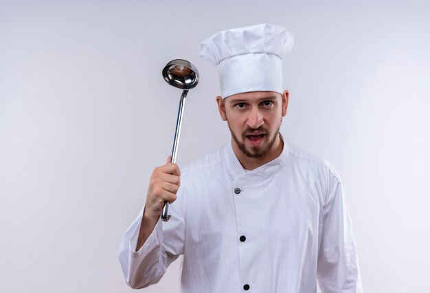 Cocinero cocinero profesional masculino en uniforme blanco y sombrero de cocinero balanceando un cucharón con cara enojada de pie sobre fondo blanco.