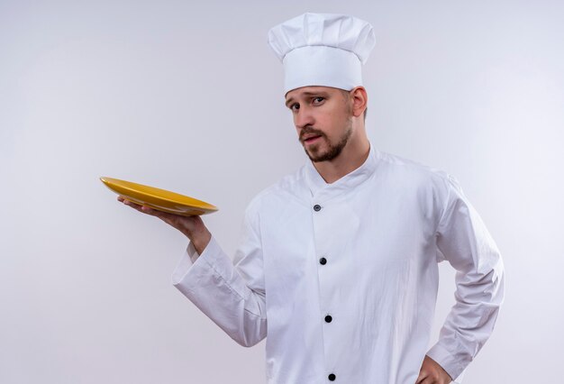 Cocinero cocinero profesional masculino en uniforme blanco y sombrero de cocinero demostrando un plato vacío con mirada confiada de pie sobre fondo blanco