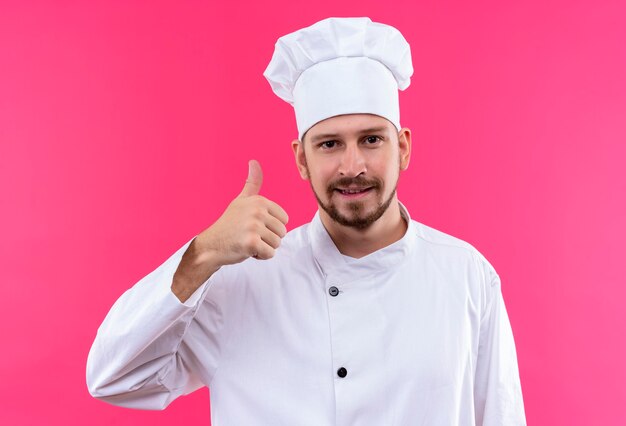 Cocinero cocinero profesional masculino en uniforme blanco y sombrero de cocinero mirando a la cámara sonriendo amigable mostrando los pulgares para arriba sobre fondo rosa