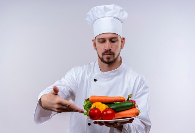 Cocinero cocinero profesional masculino en uniforme blanco y sombrero de cocinero presentando un plato con verduras mirando confiado de pie sobre fondo blanco