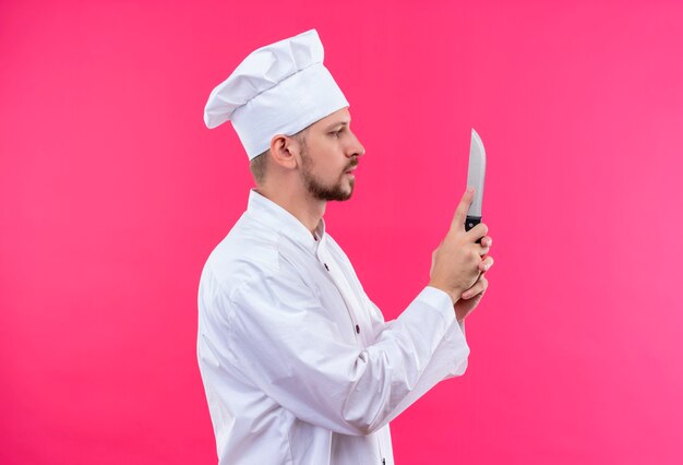 Cocinero cocinero profesional masculino en uniforme blanco y sombrero de cocinero sosteniendo un cuchillo de pie hacia los lados sobre fondo rosa