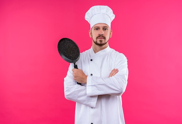 Cocinero cocinero profesional masculino en uniforme blanco y sombrero de cocinero sosteniendo una sartén con los brazos cruzados mirando confiado de pie sobre fondo rosa