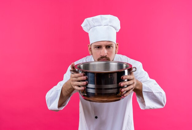 Cocinero cocinero profesional masculino en uniforme blanco y sombrero de cocinero sosteniendo una sartén vacía mirando intrigado de pie sobre fondo rosa